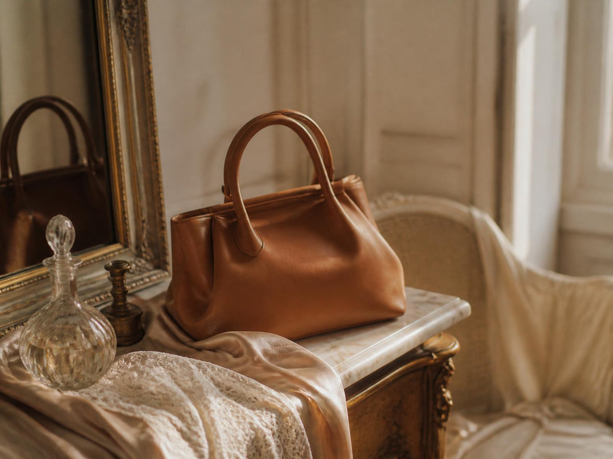 A caramel leather handbag, glass perfume decanter and ivory lace on a marble dressing table in warm daylight